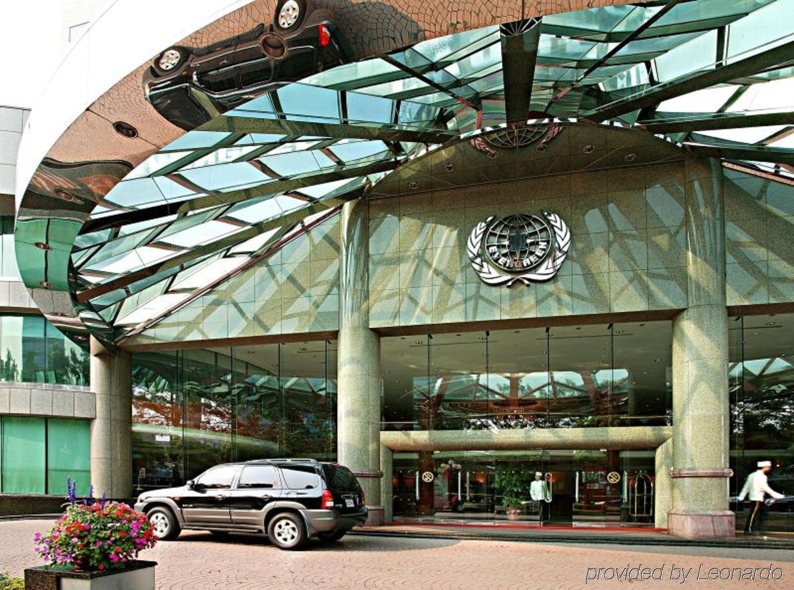 Evergreen Laurel Hotel - Тайчжун Экстерьер фото The photo shows the entrance of a modern building characterized by its glass and metal architecture. The structure features a large, domed roof that reflects light, with a distinctive emblem or logo displayed prominently at the center. In front of th