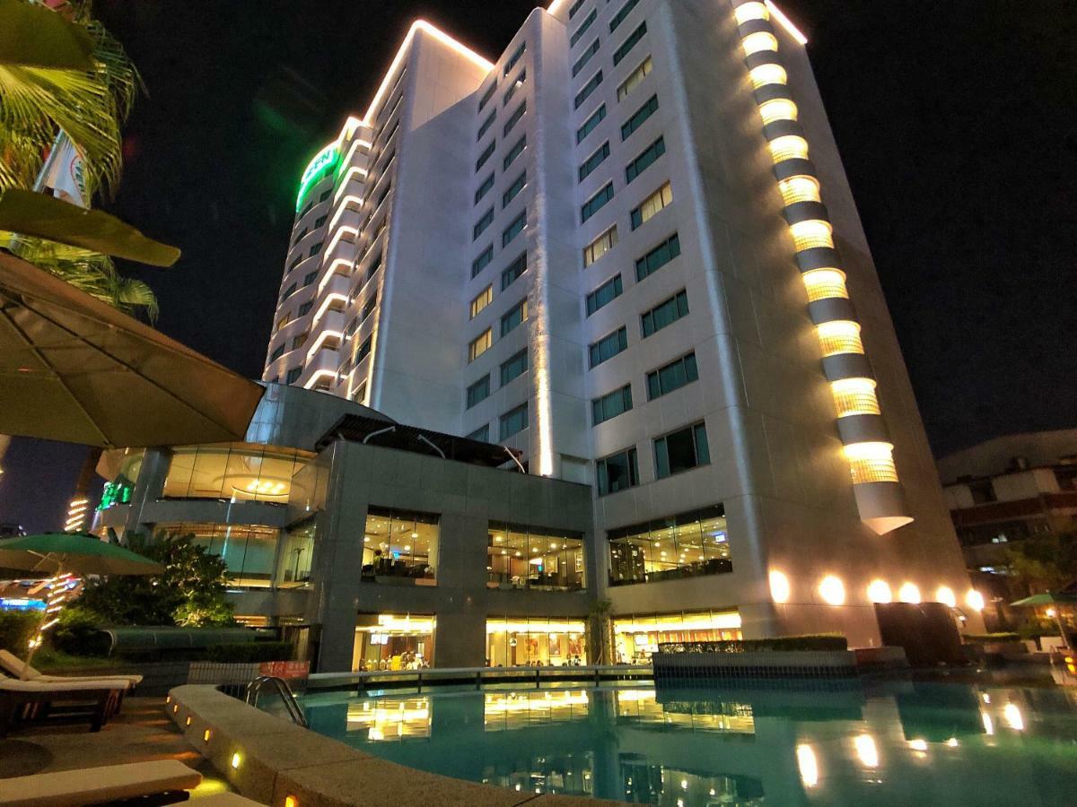 Evergreen Laurel Hotel - Тайчжун Экстерьер фото The photo shows a tall hotel building at night, illuminated by lights. The architecture appears modern, with several floors and large windows. In the foreground, there is a swimming pool that reflects the lights from the building, creating a serene a