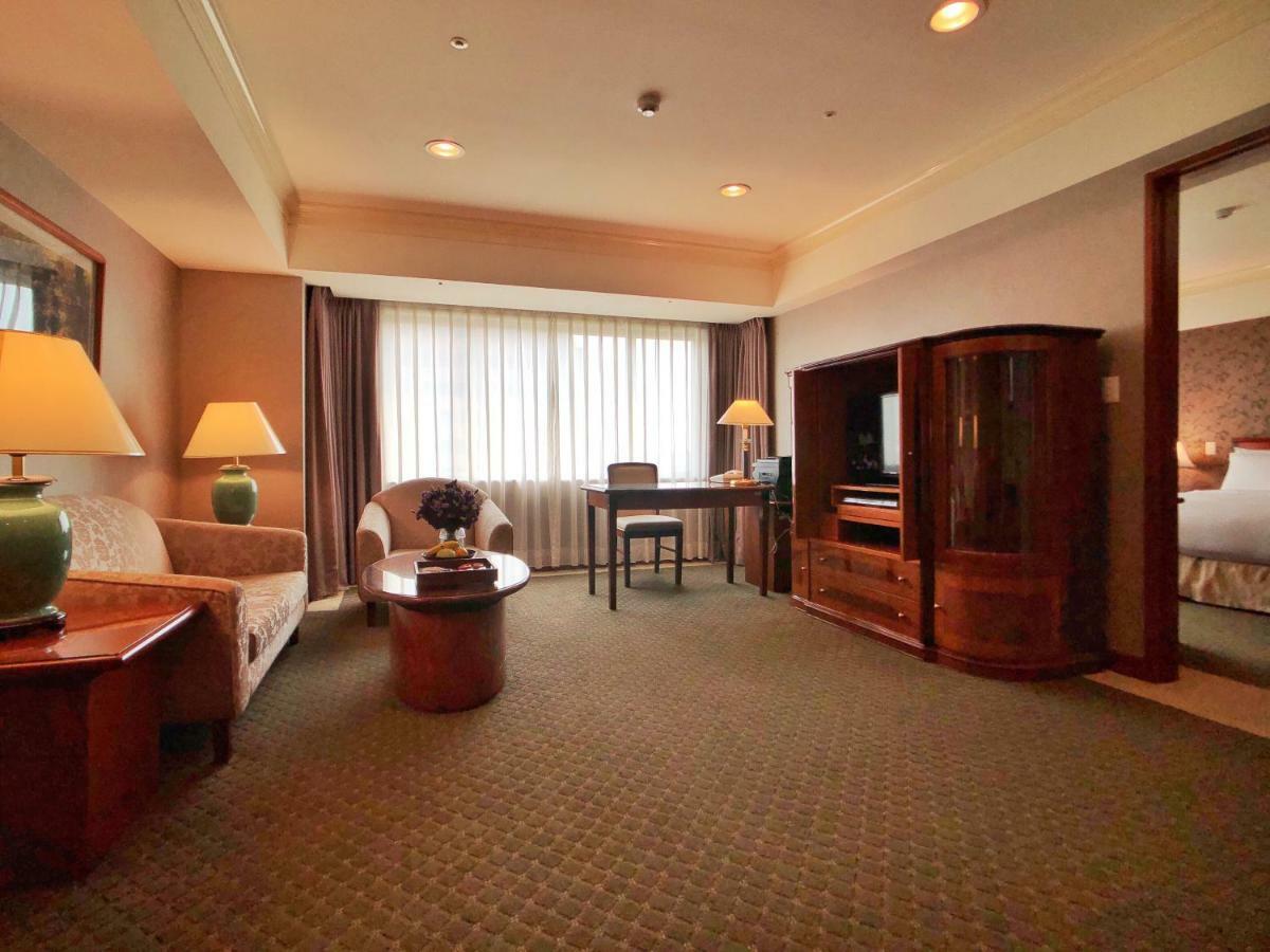 Evergreen Laurel Hotel - Тайчжун Экстерьер фото The photo shows a cozy hotel suite or living area. There is a sofa and an armchair arranged around a small coffee table. A desk with a chair is positioned near a large window covered with sheer curtains that allow natural light to enter the room. Two